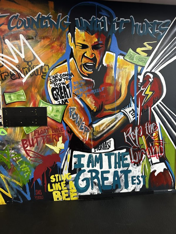 Hand-painted mural of a boxer with arms raised, surrounded by phrases including 'I AM THE GREATEST,' 'FLOAT LIKE A BUTTERFLY,' and 'STING LIKE A BEE'