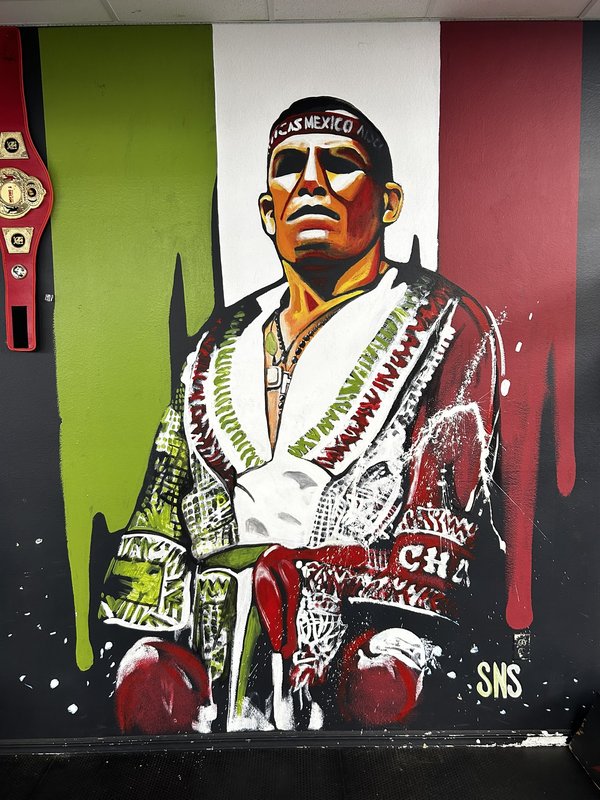 Hand-painted mural of a boxer in a championship robe, painted within the green-white-red stripes of the Mexican flag, beside a real championship belt mounted on the wall